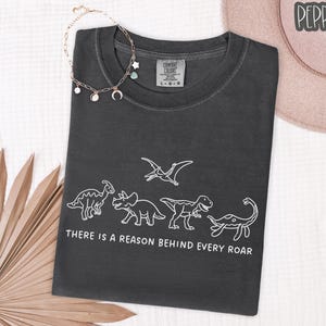 May include: A charcoal gray short-sleeve t-shirt featuring a white line art graphic of five dinosaurs and the text "THERE IS A REASON BEHIND EVERY ROAR" printed below the figures. The shirt has a crew neckline and a Comfort Colors brand label.