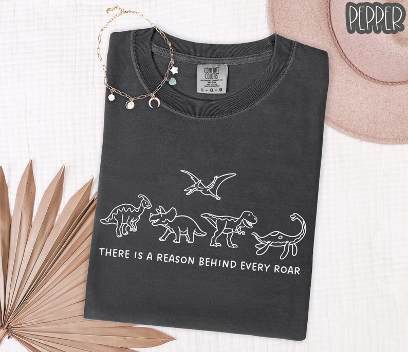 May include: A charcoal gray short-sleeve t-shirt featuring a white line art graphic of five dinosaurs and the text "THERE IS A REASON BEHIND EVERY ROAR" printed below the figures. The shirt has a crew neckline and a Comfort Colors brand label.