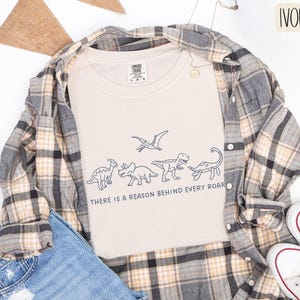 May include: An ivory t-shirt featuring a dark blue line art graphic of four dinosaurs and a pterosaur, with the text "THERE IS A REASON BEHIND EVERY ROAR" printed below. The shirt is layered under a plaid button-down shirt and blue denim jeans.