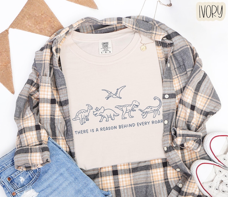 May include: An ivory t-shirt featuring a dark blue line art graphic of four dinosaurs and a pterosaur, with the text "THERE IS A REASON BEHIND EVERY ROAR" printed below. The shirt is layered under a plaid button-down shirt and blue denim jeans.
