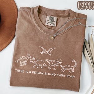 May include: An espresso-colored short-sleeve t-shirt featuring a white line-art graphic of four dinosaurs and a pterodactyl. The text below the dinosaurs reads, "THERE IS A REASON BEHIND EVERY ROAR." The shirt has a Comfort Colors brand label.