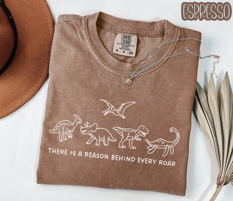 May include: An espresso-colored short-sleeve t-shirt featuring a white line-art graphic of four dinosaurs and a pterodactyl. The text below the dinosaurs reads, "THERE IS A REASON BEHIND EVERY ROAR." The shirt has a Comfort Colors brand label.