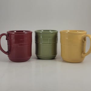 May include: Three ceramic mugs in burgundy, olive green, and yellow. The burgundy and yellow mugs have handles. Each mug has a textured, horizontal band design. The mugs are displayed on a white surface.