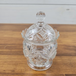 May include: A clear, vintage glass candy dish with a lid. The dish features a detailed, cut-glass pattern on the body and lid, with a decorative knob on top. The dish is sitting on a wooden surface.