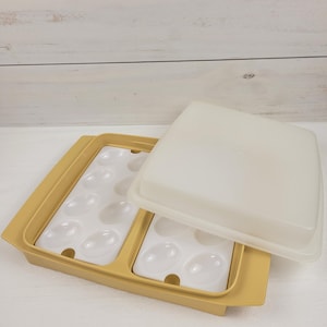 May include: A vintage egg storage container with a yellow base and a white interior. The container has a white lid and two sections with egg-shaped indentations. The container is designed to hold a dozen eggs.