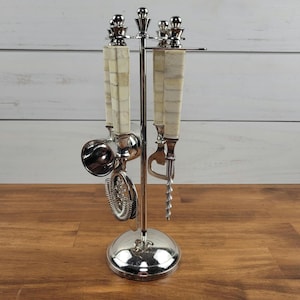May include: A silver-toned bar tool set on a circular stand featuring four utensils with cream-colored, rectangular-tiled handles. The set includes a jigger, a strainer, a bottle opener, and a corkscrew, all suspended from a central metal frame.