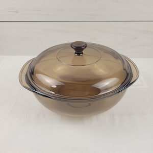 May include: A round, translucent brown glass casserole dish with integrated side handles and a matching domed glass lid featuring a central knob handle. The cookware has a smooth, polished finish and a deep, circular basin for food preparation.
