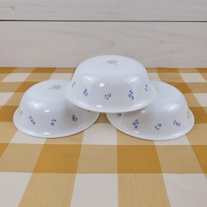 May include: Three white bowls with a rounded shape and a smooth finish. Each bowl features a delicate pattern of small blue flowers scattered across the exterior surface. The bowls are arranged in a cluster, showing their curved sides and rims.