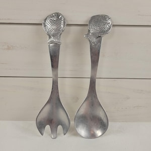 May include: A pair of silver-toned metal salad serving utensils featuring a fish-shaped handle design. One utensil is a three-pronged fork and the other is a rounded spoon. Both pieces have a textured scale pattern on the fish head handles.