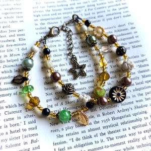 May include: A multi-strand beaded bracelet featuring a mix of yellow, green, brown, black, and white beads in various shapes, including floral and leaf motifs. The bracelet includes a bronze-toned lobster clasp and a dangling star-shaped charm.