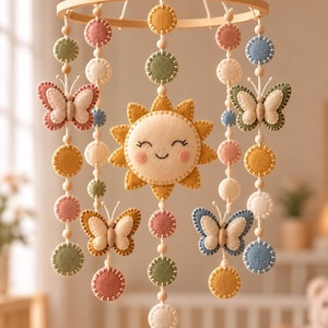 May include: A hanging mobile featuring a central felt sun with a smiling face, surrounded by felt butterflies and circular felt discs in muted shades of pink, yellow, green, and blue. The elements are suspended from a circular wooden frame.