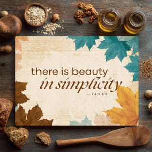 May include: A rectangular glass cutting board featuring a cream-colored background with a vintage script overlay and colorful autumn leaves in shades of teal, brown, and yellow. The text reads, there is beauty in simplicity — VAUGHN.