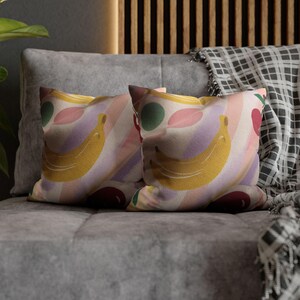 May include: Two decorative throw pillows with a colorful fruit pattern. The design features yellow bananas, red cherries, green circles, and pink leaves on a striped background. The pillows are on a gray couch.