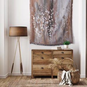 Puede incluir: Interior de una habitación con una cómoda de madera, una cesta de mimbre y una lámpara de pie. Un tapiz con un diseño abstracto marrón, blanco y gris cuelga de la pared. Una pequeña planta en maceta y libros están sobre la cómoda.