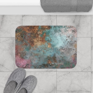 Puede incluir: Una alfombra de baño rectangular con un diseño envejecido y texturizado en tonos marrón, verde azulado y rosa. La alfombra está colocada sobre un suelo embaldosado, con un par de zapatillas grises y una toalla gris visibles.
