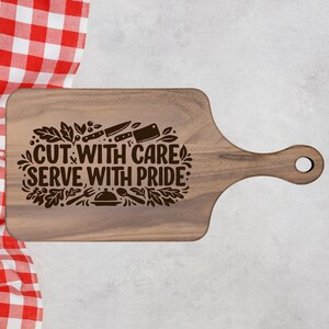 Peut inclure: Planche à découper en bois avec une poignée et un trou pour suspendre. Le texte gravé "CUT WITH CARE SERVE WITH PRIDE" est entouré d'illustrations de couteaux, de fourchettes et de feuilles. La planche est de couleur marron clair.