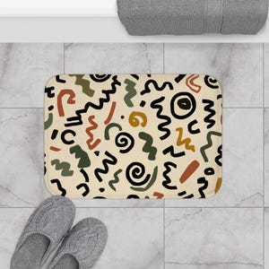 May include: A rectangular bath mat featuring a cream-colored background with a repeating pattern of black, olive green, and terracotta orange abstract shapes, including squiggly lines, spirals, and solid geometric forms across the surface.