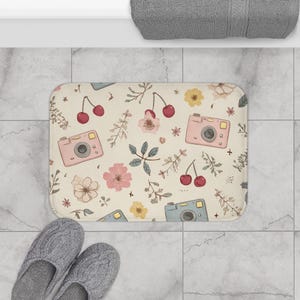 May include: A rectangular bath mat with a cream-colored background featuring a repeating pattern of pink vintage cameras, red cherries, and various small flowers and leaves in shades of yellow, pink, and muted green across the entire surface.