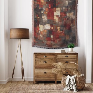 May include: A woven wall tapestry with a geometric pattern in shades of red, brown, and gray hangs above a wooden dresser. A floor lamp with a brown shade and brass legs stands to the left. A woven basket and small potted plant are on the dresser.
