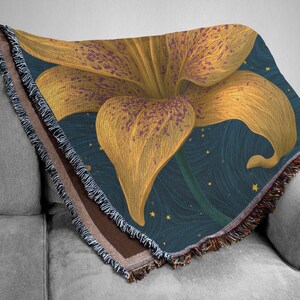 May include: A woven throw blanket featuring a large, golden-yellow lily with purple speckles centered on a dark blue background. The background displays a swirling pattern with small yellow stars. The blanket has a fringed edge with multi-colored threads.