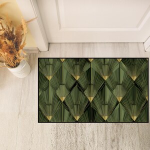 May include: A rectangular doormat with a geometric art deco design. The mat features a repeating pattern of green and gold fan shapes, outlined in black. The mat is placed on a light wood floor.