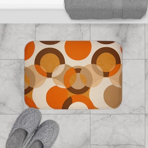May include: A rectangular bath mat with a retro design. The mat features overlapping circles in shades of orange, brown, and beige. A pair of gray knitted slippers are visible in the lower left corner. A gray towel is in the upper right corner.