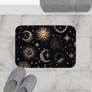 Può includere: Un tappetino da bagno rettangolare nero con un design celeste dorato, con soli, lune, stelle e costellazioni. Il tappetino è posizionato su un pavimento piastrellato, con un paio di pantofole grigie in primo piano.
