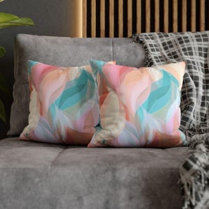 May include: Two square throw pillows featuring a repeating abstract pattern of flowing, wave-like shapes in soft shades of peach, light pink, teal, and cream. The pillows have a smooth, uniform texture and are displayed side by side on a gray surface.