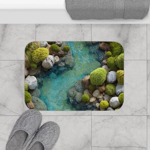 May include: A rectangular bath mat featuring a detailed design of a stream with mossy rocks and vibrant green moss. The mat has a turquoise blue water effect and is surrounded by gray rocks. The mat is placed on a tiled floor.