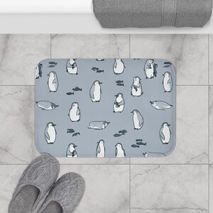 May include: A rectangular bath mat with a light blue background featuring a repeating pattern of illustrated penguins in various poses, including standing, swimming, and lying down, interspersed with small dark gray fish icons.