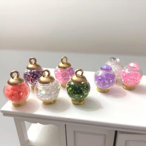 May include: A collection of small, clear glass globe charms filled with colorful glitter and sequins. Each charm has a gold-colored metal cap and base. The colors include red, purple, pink, white, and green.
