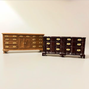 May include: Two miniature wooden dressers. One is a light brown color with gold-colored hardware. The other is a dark brown color with gold-colored hardware. Both have multiple drawers and are on short legs.