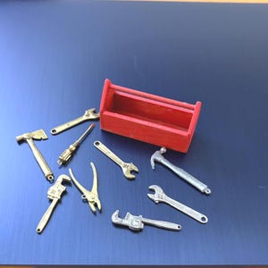 May include: A miniature red wooden toolbox accompanied by a collection of small metal tool charms, including a hatchet, several wrenches, a screwdriver, pliers, and a hammer, all arranged together as a set of decorative hardware accessories.