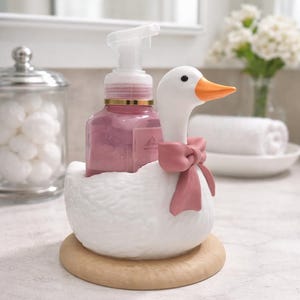 May include: A white ceramic goose-shaped soap dispenser with a pink bow, holding a pink soap bottle. The dispenser sits on a wooden base. A glass jar with cotton wool balls is in the background.