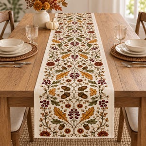 May include: A long, rectangular table runner featuring a cream-colored fabric base with a repeating botanical pattern of brown acorns, golden-yellow oak leaves, dark green foliage, and clusters of deep red berries arranged symmetrically.