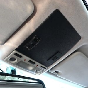 May include: Interior car roof panel with a black, textured surface. The panel features a light fixture with multiple buttons and a small vent. The sun visors are light gray.