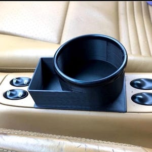 May include: A black, 3D-printed car cup holder and storage tray. The cup holder is a circular design, and the tray is rectangular. The item is placed on a tan car console with control buttons and tan leather seats in the background.