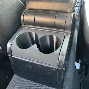 May include: Black car console with two cup holders. The console is made of plastic and is located between the front seats. The armrest is black leather. The cup holders are circular and designed to hold beverages.