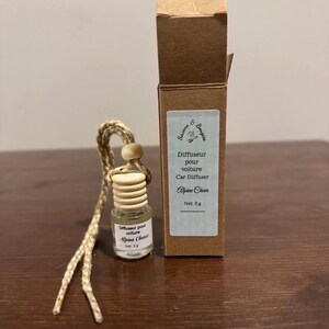 Peut inclure: Un petit diffuseur de parfum pour voiture en verre avec un bouchon en bois et une corde tressée. L'étiquette indique "Alpine Cheer". Une boîte en carton avec l'inscription "Diffuseur pour voiture Car Diffuser Alpine Cheer Net 5 g" est à côté.