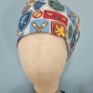 May include: A light blue scrub cap featuring various colorful emblems and symbols. The cap is adorned with images of a golden snitch, crossed wands, and house crests. The cap is displayed on a mannequin head against a light blue background.