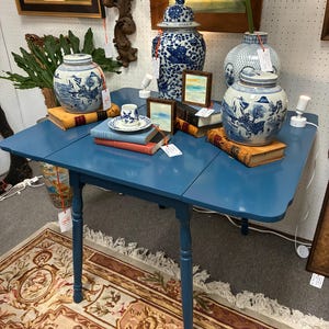 May include: A blue drop-leaf table with four slender legs, displaying three blue and white ceramic ginger jars, a small teacup on a saucer, two framed landscape paintings, and several vintage books stacked on the tabletop surface.