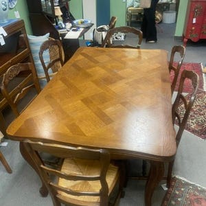 May include: A rectangular wooden dining table with a parquet pattern, surrounded by six wooden chairs with woven seats. The table and chairs are a warm, medium brown colour, suggesting a vintage or antique style. The chairs have curved backs.