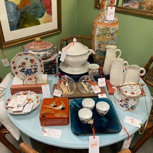 Puede incluir: Una colección de artículos de cerámica decorativos, que incluyen un jarrón grande, una sopera, platos, tazas de té y una tetera, dispuestos sobre una mesa redonda azul claro. Los artículos presentan diseños florales y paisajísticos.