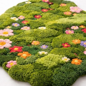 Könnte beinhalten: Ein grüner, strukturierter Teppich mit floralem Design. Der Teppich weist verschiedene Grüntöne auf, mit bunten, gestickten Blumen in Rosa, Orange, Rot, Gelb und Lila. Der Teppich hat eine unregelmäßige, organische Form.
