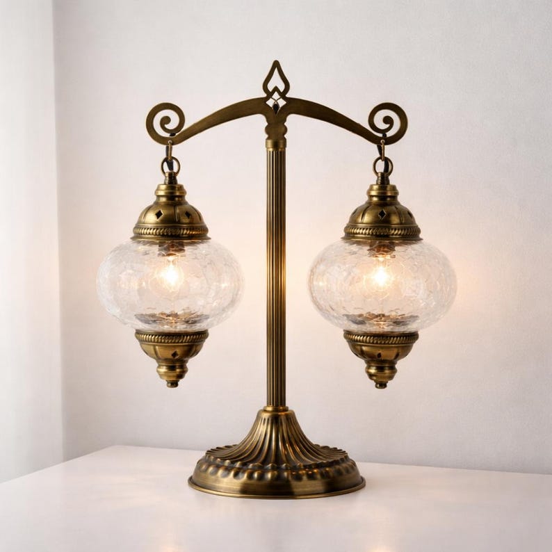 May include: An antique brass table lamp featuring two clear, crackled glass globes. The lamp has a central support with a decorative top and base. The globes are suspended from curved arms. The lamp is lit, emitting a warm glow.