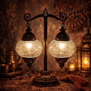 May include: A decorative table lamp featuring two spherical, crackled glass shades. The lamp has a bronze-coloured metal base and a curved top. The shades are illuminated, emitting a warm light. The lamp is designed in an ornate, vintage style.