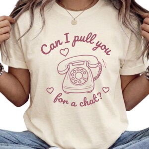 May include: A cream-colored short-sleeve t-shirt featuring a maroon graphic of a rotary telephone. The text "Can I pull you for a chat?" is printed in maroon above and below the telephone, accompanied by three small maroon heart icons.
