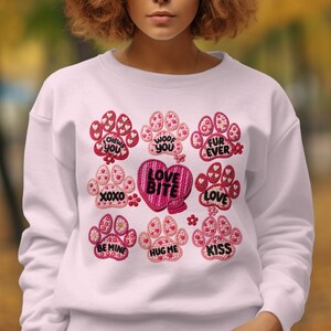 May include: White sweatshirt featuring embroidered paw prints in pink and red, with phrases such as "Chews You", "Woof You", and "Love". A heart with "Love Bite" is in the center. A fun and cute design.