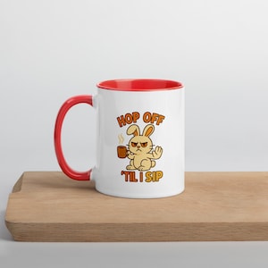 May include: White ceramic mug with a red interior and handle. The mug features a cartoon rabbit with an angry expression holding a coffee cup. Text on the mug reads "HOP OFF 'TIL I SIP".
