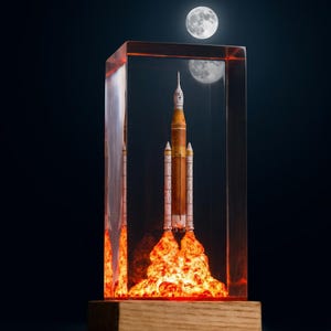 May include: A detailed model rocket launching, encased in a clear rectangular block. The rocket is orange and white, with flames at the base. A full moon is visible in the background, creating a space theme.
